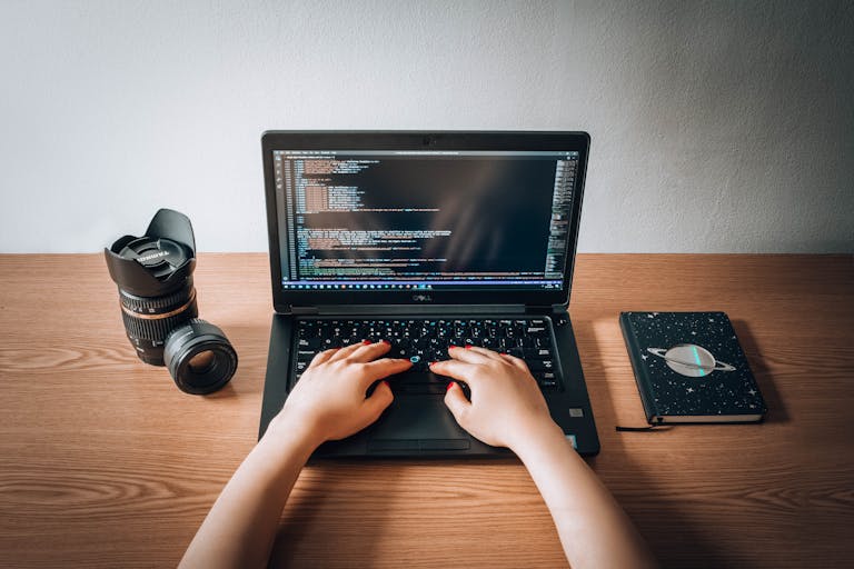 A woman coding on a laptop with a camera lens and notebook on a wooden desk in a home office.